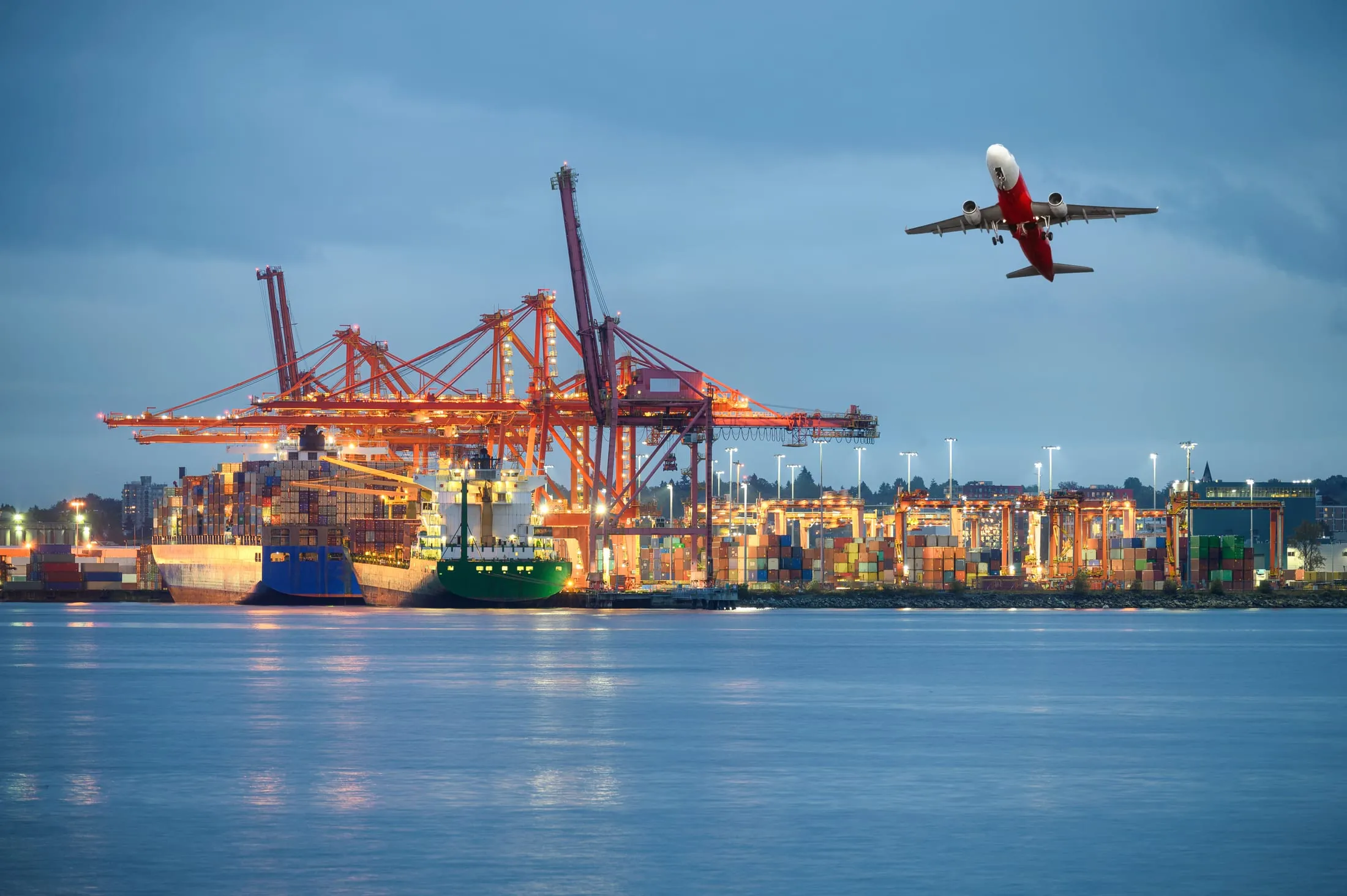 Avión despegando sobre las grúas y buques de un puerto de contenedores al atardecer